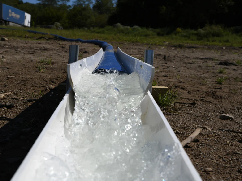 A close-up view of clean, clear water flowing forcefully from a blue discharge pipe into a white V-shaped metal channel on bare ground, with grass visible in the background.