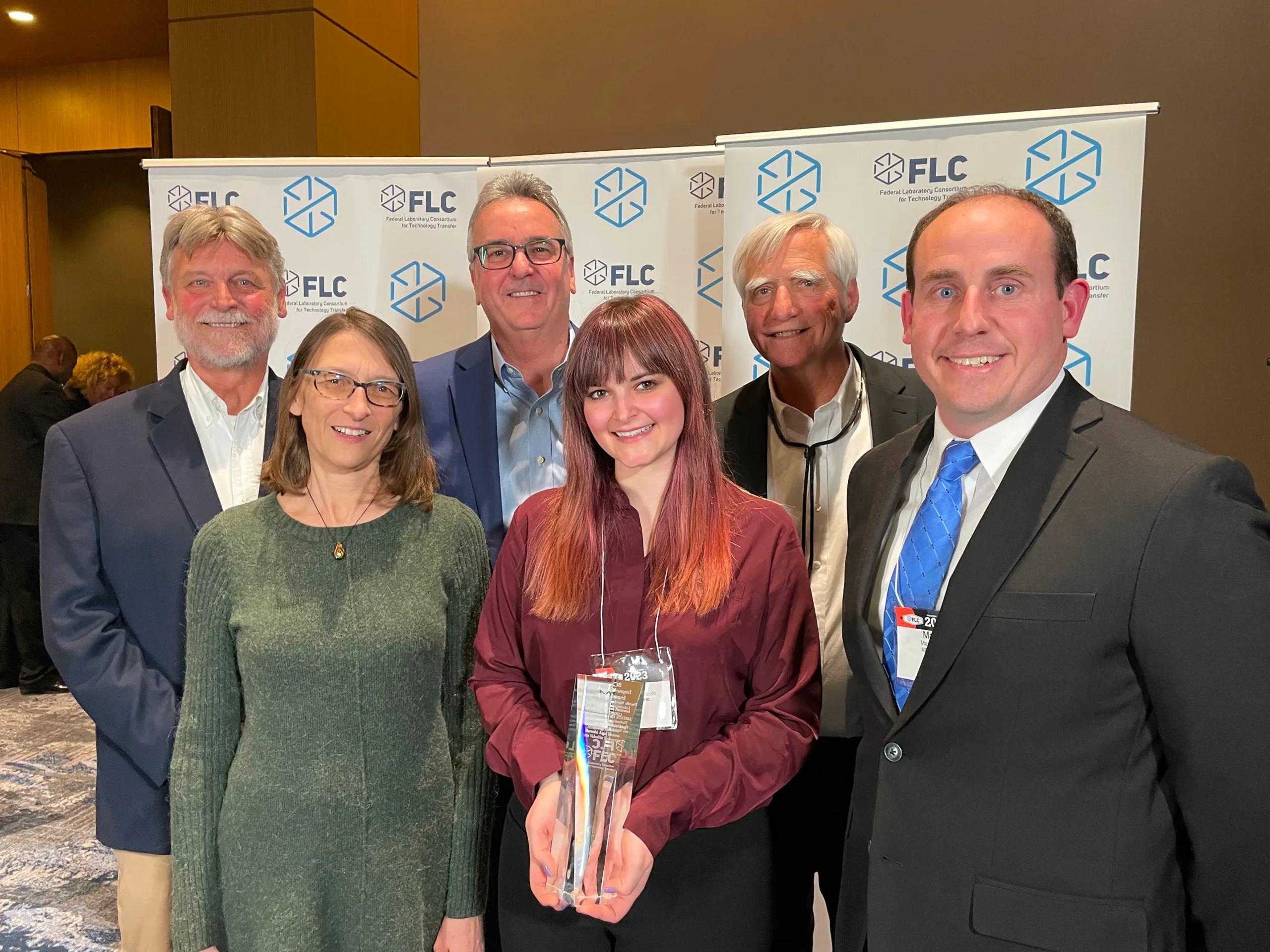 A group of six BlueCycle team members smiling at an awards event, with one person holding a 2023 FLC award trophy in front of Federal Laboratory Consortium for Technology Transfer branded banners.