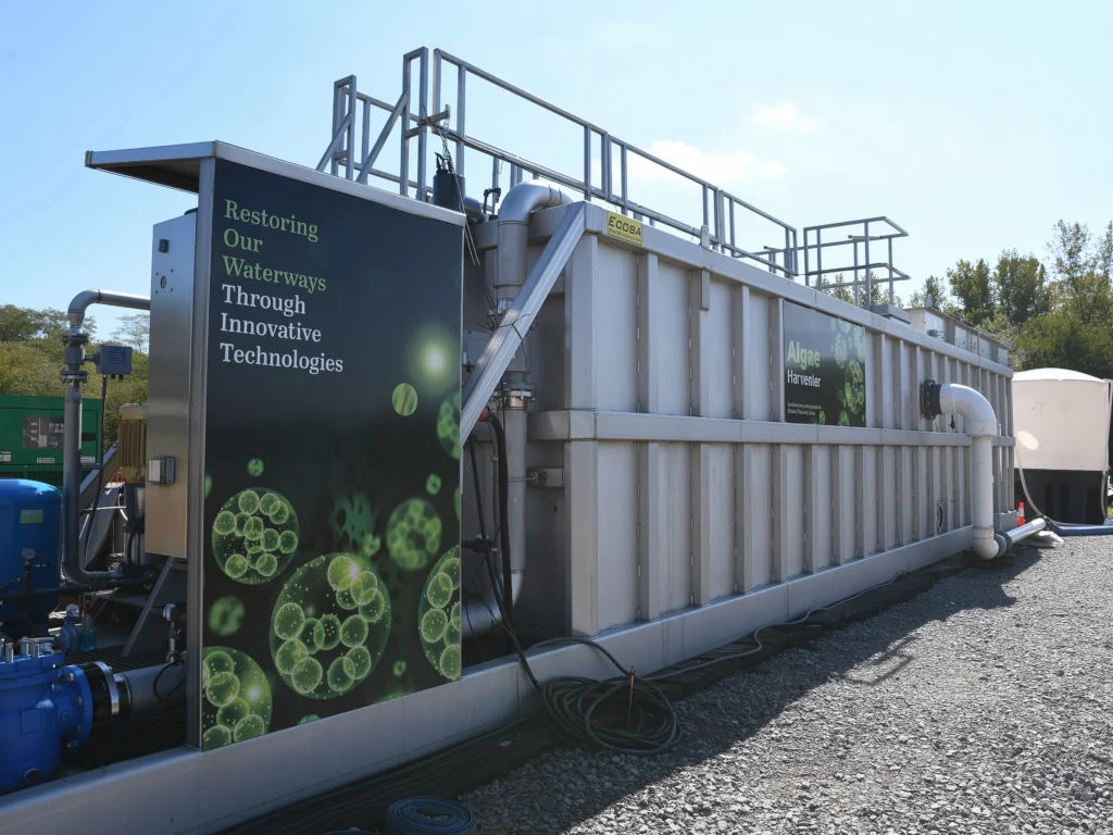 Large-scale algae harvester unit deployed outdoors, with branded panels showing microscopic algae imagery