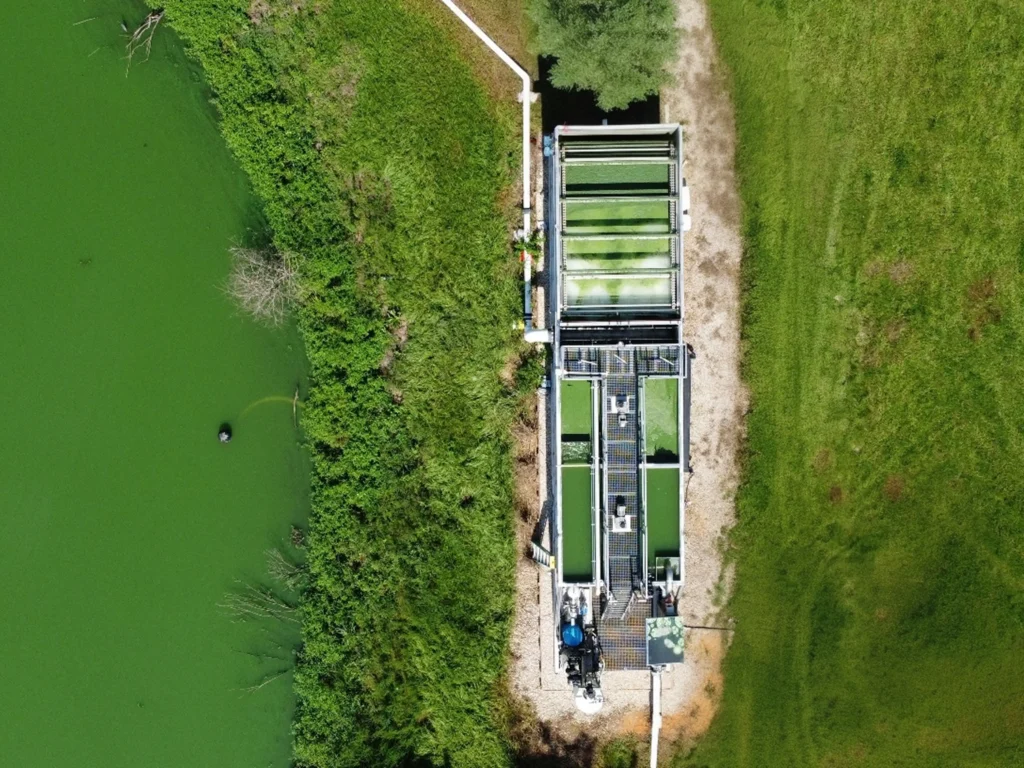 Aerial view of an HFT™ algae harvesting system positioned at the edge of a heavily algae-bloomed green waterway, with processing tanks and piping visible alongside a gravel access path and surrounding grassland.
