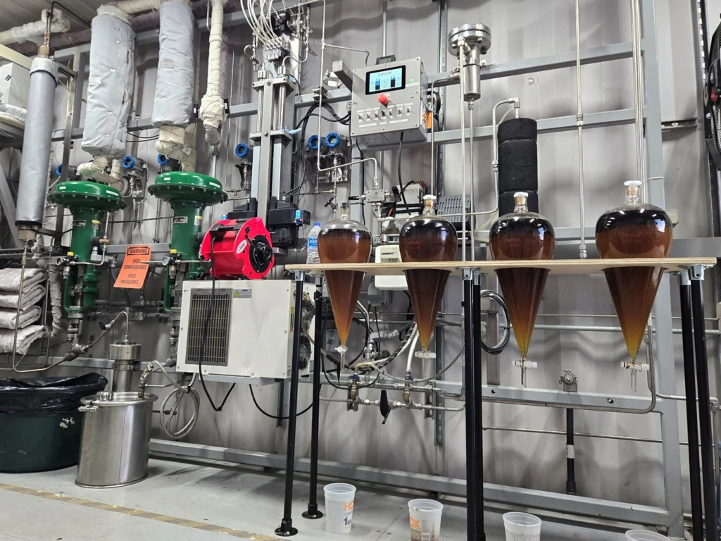 Interior view of an HTL-360™ hydrothermal liquefaction facility showing four conical glass separatory funnels containing dark amber biocrude oil, surrounded by high-pressure industrial piping, pumps, and control equipment.